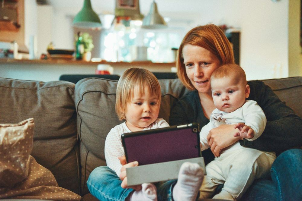A mother holding her baby while her toddler looks at a tablet on the sofa